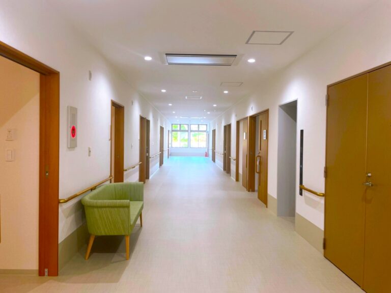 介護施設の廊下。複数の居室ドアと明るい照明、共用ソファが配置されている。
